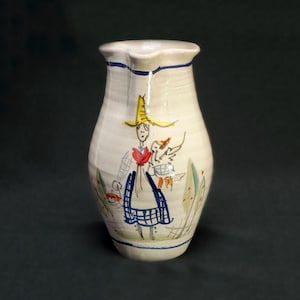 May include: Cream-colored ceramic pitcher with a hand-painted illustration of a person in a yellow hat and blue apron carrying a basket. The pitcher has a ruffled rim and blue trim. Decorative leaves and flowers are painted around the figure.