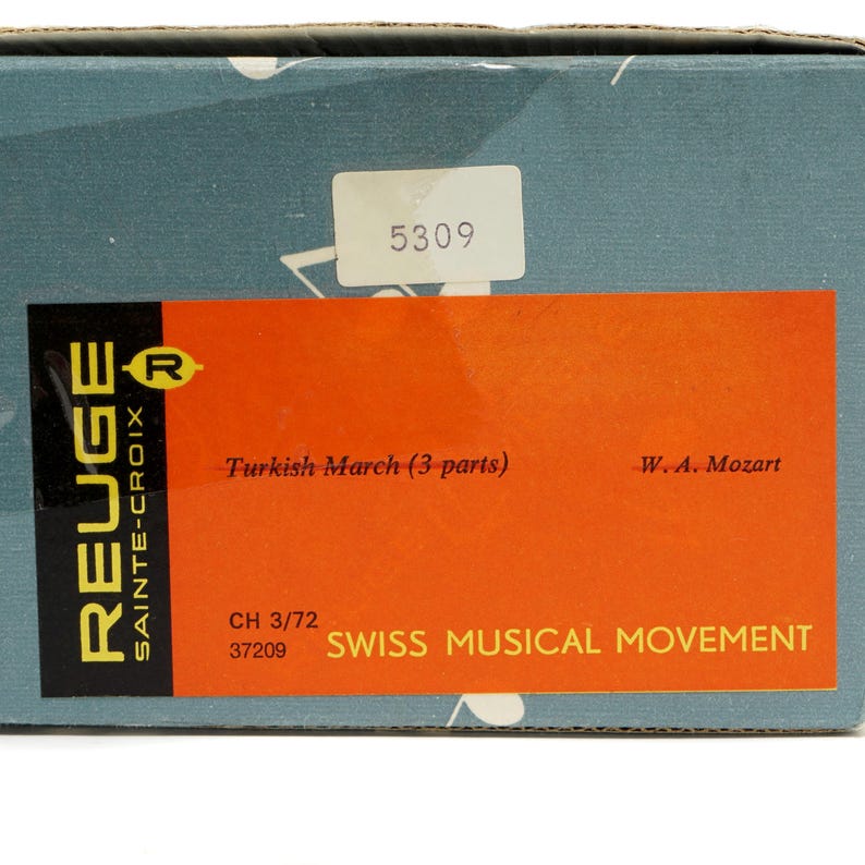 May include: A close-up of a box with a label that reads "Turkish March (3 parts)" by W.A. Mozart. The label is orange and has the text "SWISS MUSICAL MOVEMENT". The box is teal with the brand name "REUGE" in white.