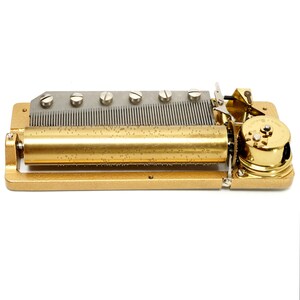 May include: A vintage gold-toned music box mechanism. The mechanism features a rotating cylinder with pins, a comb-like structure, and a small clockwork mechanism. The rectangular base is also gold-toned. The music box is designed to play a melody.