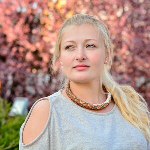 以下が含まれることがあります： 長いブロンドの髪の女性は、肩がカットアウトされたグレーのセーターを着ています。彼女はカラフルなビーズネックレスをしています。