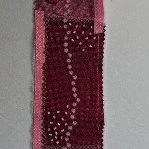May include: A handmade textile bookmark in shades of pink and burgundy. It features layered fabrics, embroidered details, beads, and decorative buttons. The top is rolled, showing a layered interior. The bookmark is approximately 25 cm long.