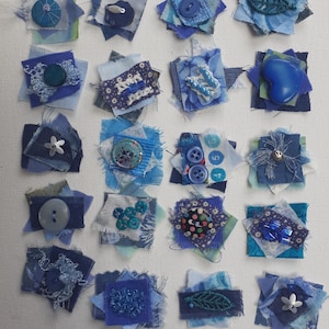May include: A collection of twenty-one small, square fabric patches in shades of blue. Each patch is decorated with buttons, beads, and other embellishments. The patches are arranged on a white surface.