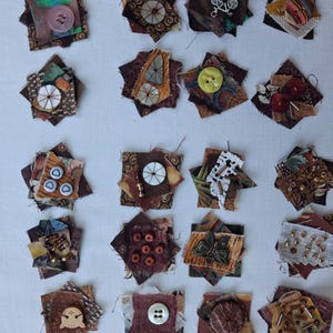 May include: A collection of twenty square fabric patches arranged in a grid, each featuring layered textile scraps in shades of brown, tan, and rust, embellished with various buttons, metal charms, beads, and decorative stitching in diverse patterns.