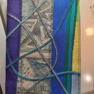 May include: A handmade notebook cover with a patchwork design. The cover is made of different fabrics in shades of blue, green, purple, and grey. The fabrics are sewn together with decorative stitching.