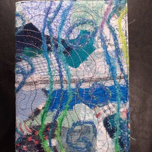 May include: A handmade journal cover featuring a blue, green and white patchwork design. The cover is decorated with stitched lines and swirls.