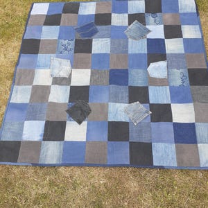 May include: A patchwork quilt made from squares of denim in various shades of blue, grey, and black. Several denim pockets are sewn onto the quilt. The quilt is laid out on a grassy surface.