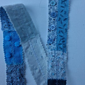 May include: A blue textile ribbon, crafted from assorted fabric pieces. The ribbon is adorned with buttons, beads, and intricate stitching. The design incorporates floral and heart motifs, resulting in a textured and detailed craft item, suitable for various projects.