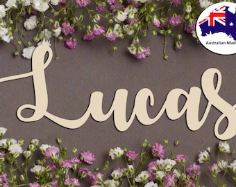 May include: Wooden name sign with the name "Lucas" written in a cursive script. The sign is made of natural wood and has a light brown color. The sign is surrounded by white and pink flowers.