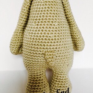 May include: A crocheted stuffed animal in the shape of a bear. The bear is wearing a pink and brown crocheted hat. The bear is standing on a white surface. The text "At Stitches End" is written on the surface.