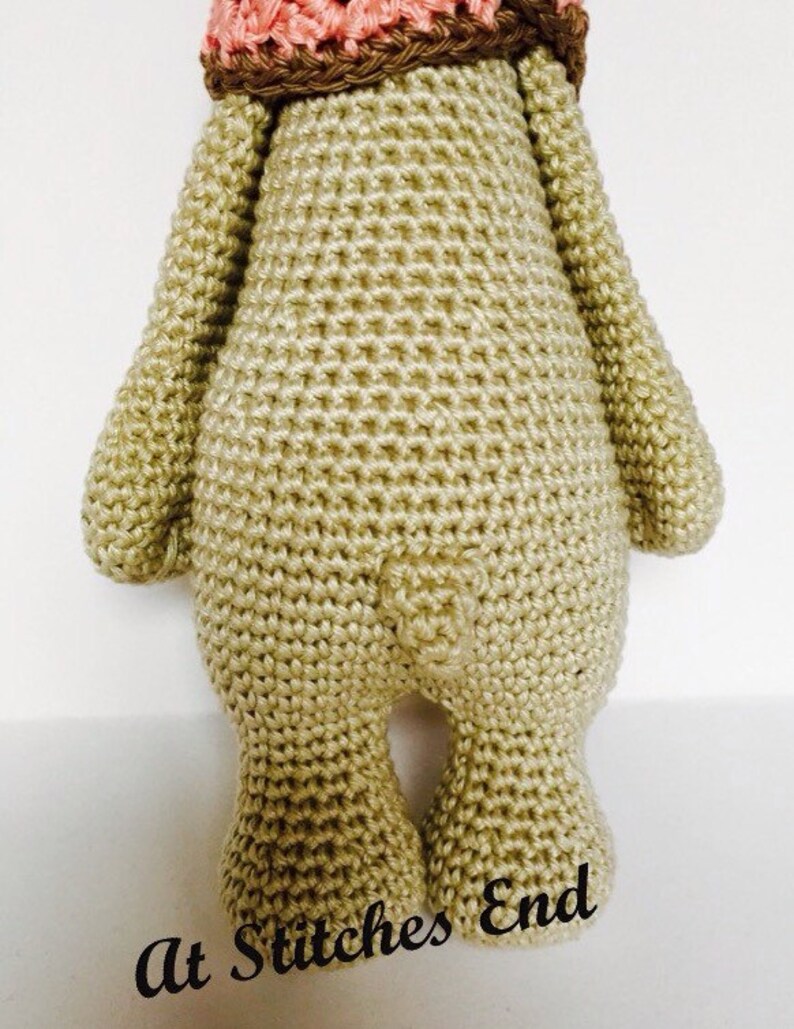 May include: A crocheted stuffed animal in the shape of a bear. The bear is wearing a pink and brown crocheted hat. The bear is standing on a white surface. The text "At Stitches End" is written on the surface.