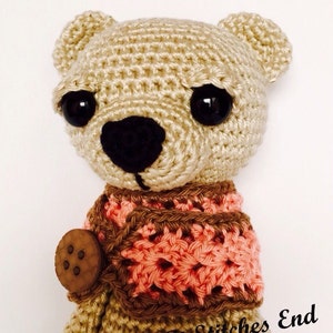 May include: A crocheted beige bear with black eyes and a brown and pink crocheted scarf. The bear has a wooden button on its chest. The bear is a handmade toy.