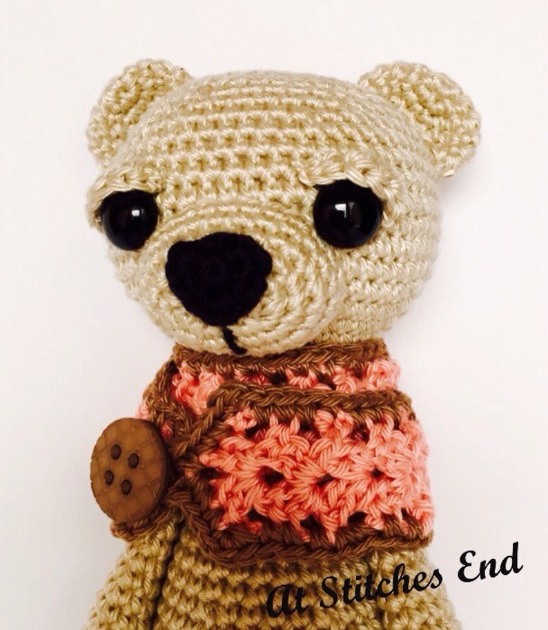 May include: A crocheted beige bear with black eyes and a brown and pink crocheted scarf. The bear has a wooden button on its chest. The bear is a handmade toy.