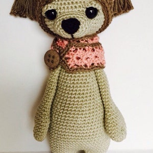 May include: A crocheted stuffed animal wearing a pink hat and scarf. The animal has brown eyes, a brown nose, and a beige body. The text "At Stitches End" is visible at the bottom of the image.