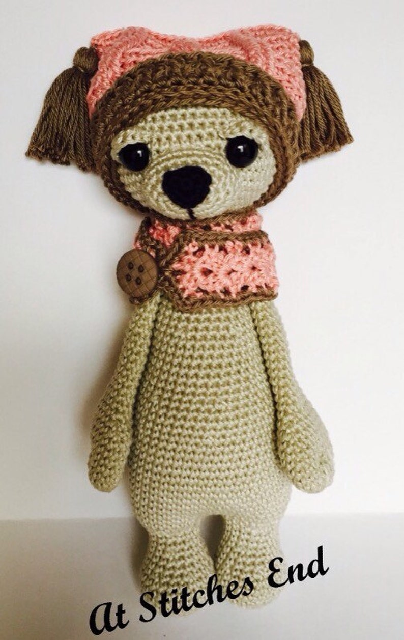 May include: A crocheted stuffed animal wearing a pink hat and scarf. The animal has brown eyes, a brown nose, and a beige body. The text "At Stitches End" is visible at the bottom of the image.