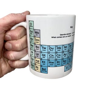 May include: White ceramic mug with a periodic table design featuring Geordie slang words instead of chemical elements. The mug has a blue, light blue, and yellow colour scheme. The text on the mug reads "Geordie element What comes oot ya mooth".
