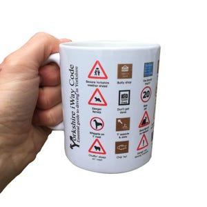 May include: White ceramic mug with a black and red design featuring a Yorkshire iWay Code. The mug has a variety of road signs with humorous Yorkshire-themed warnings, such as "Beware Yorkshire weather ahead", "Danger ferrets", "Whippits on t' road", and "Chuffin' sheep on' road".