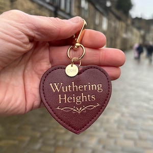 May include: A burgundy heart-shaped keyring with the words "Wuthering Heights" in gold lettering. The keyring has a gold-coloured clasp and a small circular tag with the initials "COH".