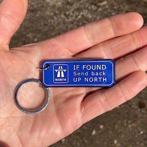 May include: A blue rectangular keyring with a silver ring. The keyring features white text: "IF FOUND Send back UP NORTH" and a highway sign graphic with the word "NORTH".