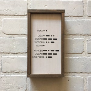 Könnte beinhalten: Ein Schild in einem Holzrahmen mit weißem Hintergrund und schwarzem Text. Das Schild zeigt das NATO-phonetische Alphabet in Morsecode. Die Wörter auf dem Schild sind: INDIA, LIMA, OSCAR, VICTOR, ECHO, YANKEE, OSCAR, UNIFORM.