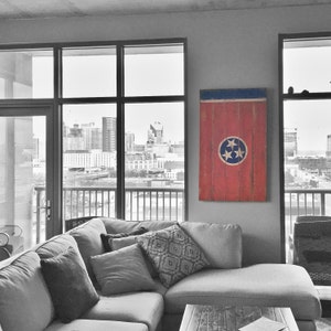 May include: A gray sectional sofa with a red, white, and blue Tennessee flag hanging on the wall behind it. The sofa is in a modern living room with large windows overlooking a city skyline.