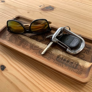 May include: A wooden tray with a topographic map design and the word "ALLEN" engraved on the bottom. The tray contains a pair of sunglasses and a key with a carabiner.