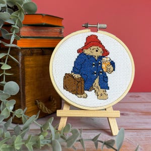 May include: A cross-stitch embroidery of a bear in a blue coat and red hat, holding a suitcase and a mug. The embroidery is in a wooden hoop on a small wooden easel. The background is a red wall.
