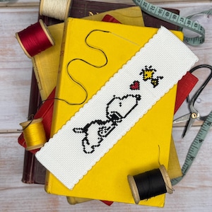 May include: A white, cross-stitched bookmark featuring Snoopy and Woodstock with a heart. The bookmark is placed on a stack of books, with spools of thread and sewing tools in the background. The bookmark has a scalloped edge.