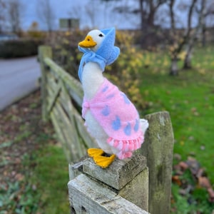 Può includere: Un'oca di feltro bianco che indossa una sciarpa con cappuccio blu e rosa. L'oca è appollaiata su un palo di legno di una recinzione.