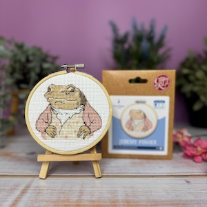 May include: A cross-stitch embroidery hoop featuring a frog wearing a pink jacket and a white shirt. The hoop is on a wooden easel and the packaging is visible behind it. The packaging says "Jeremy Fisher".