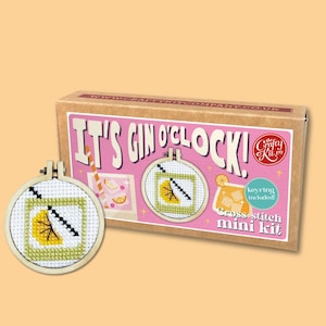 Pode incluir: Um mini kit de ponto cruz com o texto "IT'S GIN O'CLOCK!" na caixa e um desenho de cocktail em ponto cruz acabado. O kit inclui um porta-chaves. O desenho apresenta uma fatia de limão amarela e uma palhinha preta.