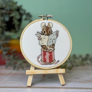 May include: A cross-stitch embroidery of a brown mouse reading a newspaper, set in a round wooden hoop. The mouse is inside a red and white striped container, displayed on a small wooden easel.