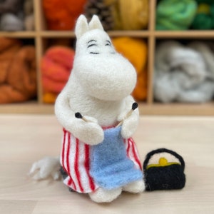 Könnte beinhalten: Ein weißes Filzspielzeug mit rot-weiß gestreiften Hosen, das einen blauen Schal mit zwei Stricknadeln strickt. Das Spielzeug hat eine schwarze Filztasche daneben.