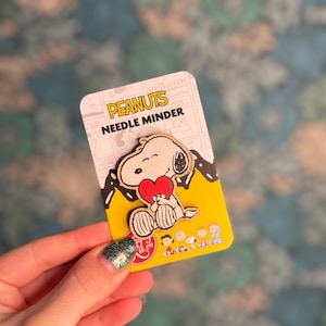 May include: A Snoopy needle minder featuring Snoopy holding a red heart. The needle minder is on a yellow and white card with the text "PEANUTS NEEDLE MINDER". The card also features illustrations of Snoopy and other Peanuts characters.