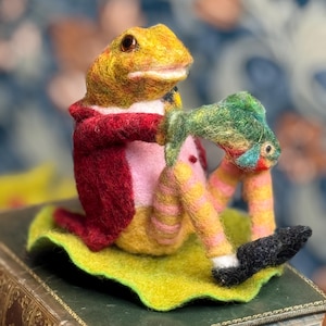 Könnte beinhalten: Eine skurrile Froschfigur aus Filz, bekleidet mit einem roten Mantel und gestreiften Hosen, die einen grünen Fisch hält. Der Frosch sitzt auf einem grünen Blatt, mit schwarzen Schuhen. Der Froschkörper ist gelb und der Fisch hat bunte Akzente.
