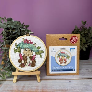 Op de afbeelding: Een kruissteekset met Benjamin Bunny uit Peter Rabbit. De set bevat een voorgedrukt Aida-doek, borduurgaren, een naald en instructies. Het afgewerkte stuk is een kleine, ronde borduurring met een kleurrijk beeld van Benjamin Bunny die twee wortels vasthoudt.