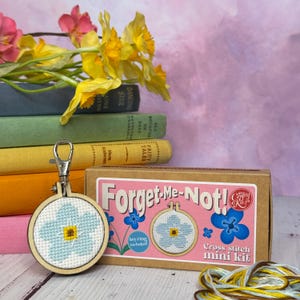 Op de afbeelding: Een mini kruissteekset met een vergeet-mij-nietje ontwerp. De set bevat een kleine houten borduurring met een blauw en geel bloemmotief, een sleutelhanger en borduurgaren. De verpakking is een bruine doos met de tekst "Forget-Me-Not!".