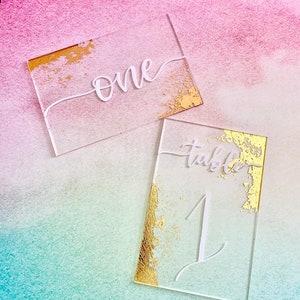 May include: Two clear acrylic table number cards with gold leaf accents. One card reads "one" in white script, the other reads "table 1". The cards are rectangular and have a modern, elegant design.