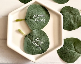 May include: Two green eucalyptus leaves with white calligraphy on a white hexagonal plate. The leaves are on a white wooden surface. The text on the leaves reads "Moon Franz" and "It is Khan".