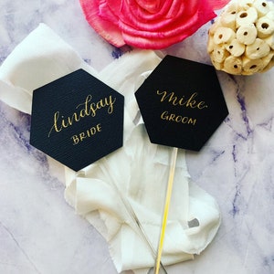 May include: Two black hexagonal signs with gold lettering on sticks. The signs say "Lindsay Bride" and "Mike Groom".