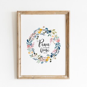Op de afbeelding: Een ingelijst print met een aquarel bloemenkrans en de tekst "Vrede zij met dit huis" met de bijbelverwijzing Lukas 10:5.