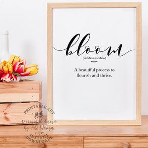 May include: A printable art print with a white background and black text. The text reads "bloom" with the definition "A beautiful process to flourish and thrive." The print is framed in a light wood frame and is sitting on a wooden table.