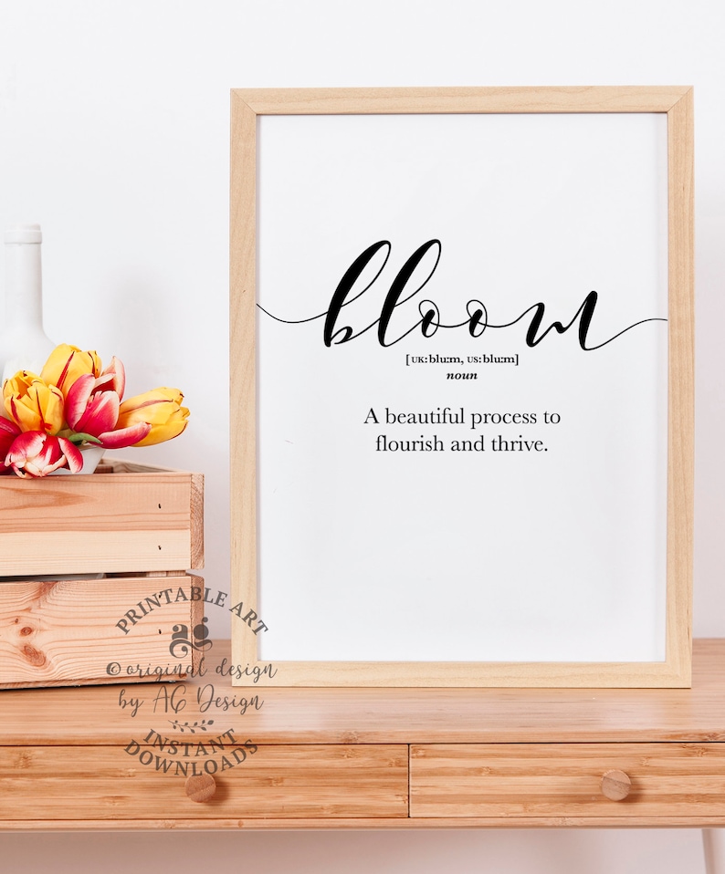 May include: A printable art print with a white background and black text. The text reads "bloom" with the definition "A beautiful process to flourish and thrive." The print is framed in a light wood frame and is sitting on a wooden table.