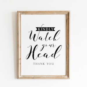 Op de afbeelding: Een ingelijst bord met zwarte kalligrafie waarop staat "Kindly Watch Your Head" met daaronder de woorden "Thank You". Het bord is in een licht houten frame.