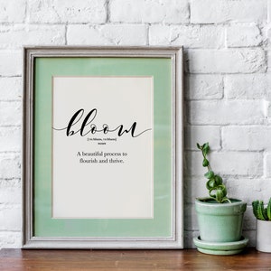 May include: A framed print with a green mat and a white background. The print features the word "bloom" in black cursive script, followed by the definition: "[bloom,  bloom] noun A beautiful process to flourish and thrive."
