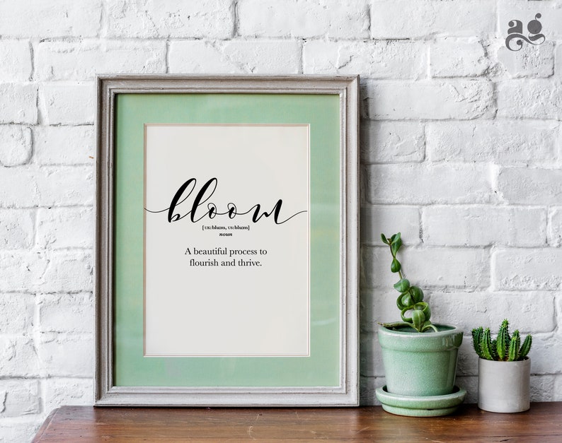 May include: A framed print with a green mat and a white background. The print features the word "bloom" in black cursive script, followed by the definition: "[bloom,  bloom] noun A beautiful process to flourish and thrive."