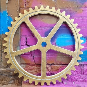 May include: A large gold-coloured gear with five spokes, set against a brick wall with pink, blue and purple spray paint.