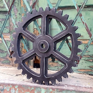 May include: A large black metal gear with six spokes. The gear is made of a rough, textured material and has a distressed look. The gear is set against a backdrop of a weathered green wooden surface.