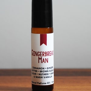May include: A roll-on perfume bottle with a black cap and a white label. The label reads "GINGERBREAD MAN" with a list of ingredients. The bottle is filled with a light brown liquid, and the background is a light gray.