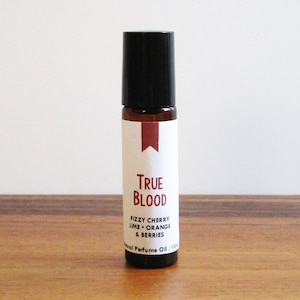 May include: A small brown glass bottle with a black roll-on top. The label is white with red text that reads "True Blood" and "fizzy cherry lime + orange & berries". The label also says "Natural Perfume Oil".