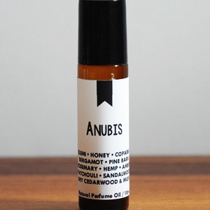 May include: A small brown glass bottle with a black roll-on top. The label is white with black text that reads "Anubis" and lists the ingredients: "Elm, Honey, Copaiba, Bergamot, Pine Bark, Rosemary, Hemp, Amber, Patchouli, Sandalwood, Dry Cedarwood & Musk". The label also says "Natural Perfume Oil | 10 ml".